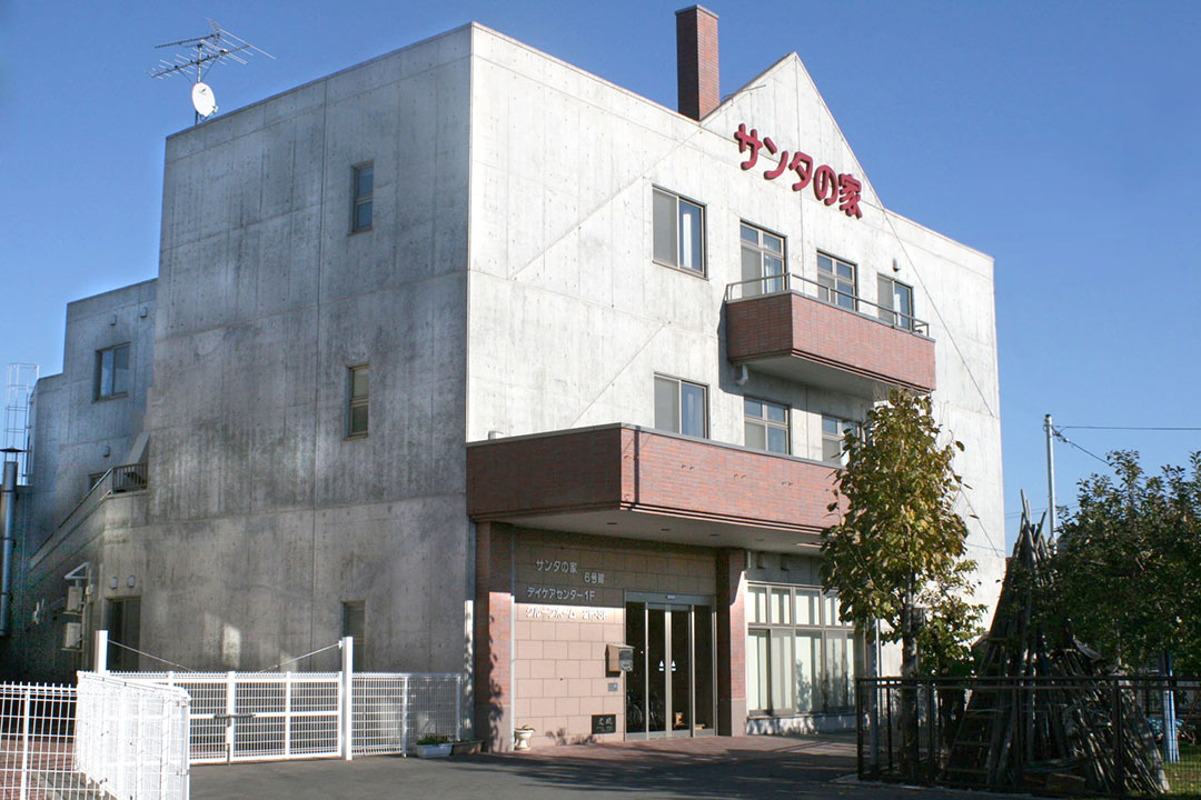 白とピンク色の3階建て介護施設の外観。青空の下、玄関と窓が見える。
